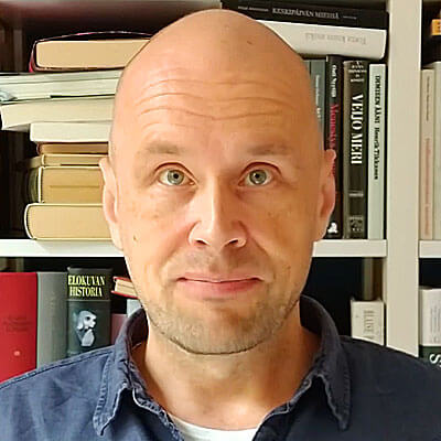 Aleksi Salokannel. There is a bookshelf filled with a diverse range of books and in front of it, a middle-aged white man looking at the camera. Bald. Not wearing a black turtleneck, however.